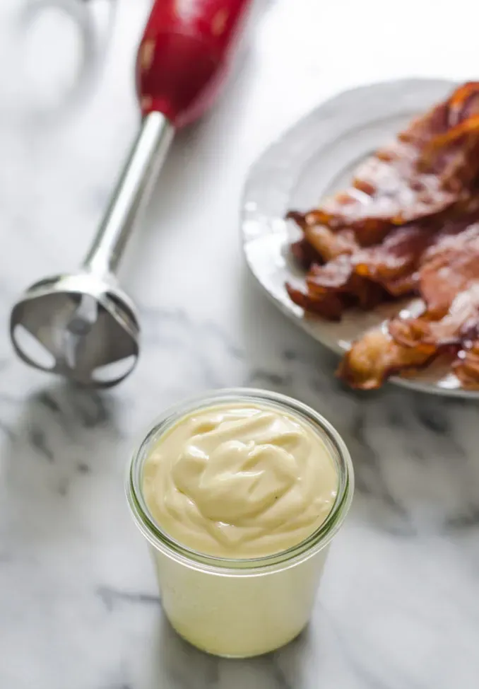 A jar of bacon fat mayo on a marble surface with a plate of cooked bacon and an immersion blender behind it.' title='Baconnaise!