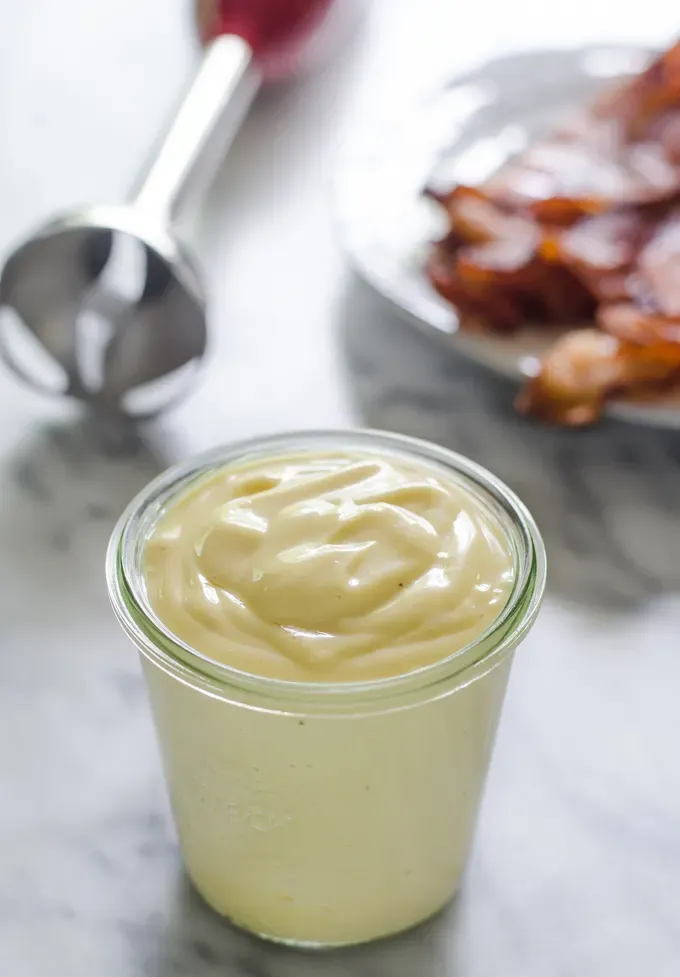 A jar of bacon grease mayonnaise on a marble surface with a plate of cooked bacon behind it.' title='Baconnaise!
