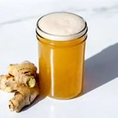 Ginger beer with foamy head, in small tumbler glass seated next to knob of fresh ginger