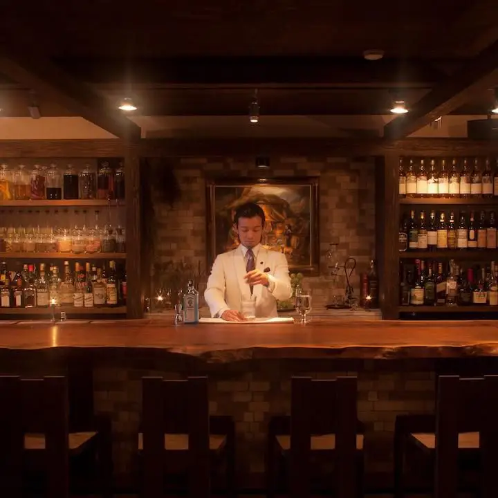 Bar Benfiddich in Tokyo. A lone bartender stands in the center of the frame, behind a abr. he is in a white blazer and is stirring a cocktail.