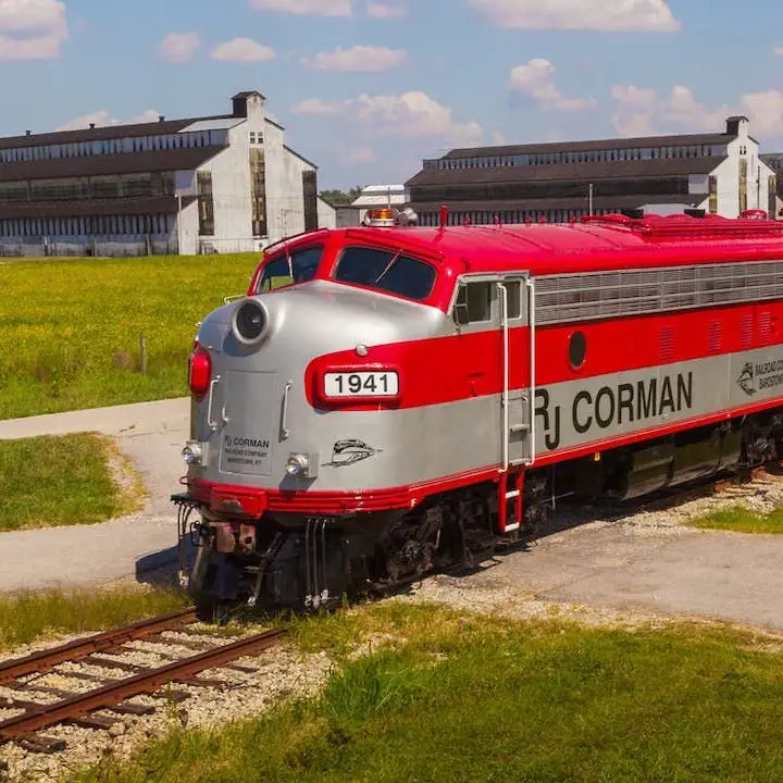 my old kentucky dinner train bourbon experience. a red and grey striped engine of a train in the foreground and distillery buildings in the background