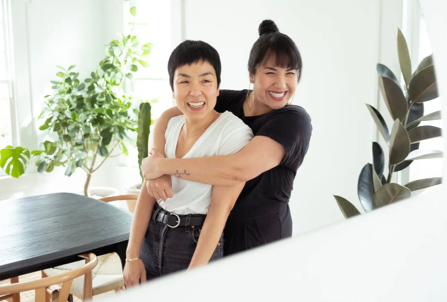 Yenvy Pham hugging Quynh-Vy Pham, both smiling, in white-walled room with plants