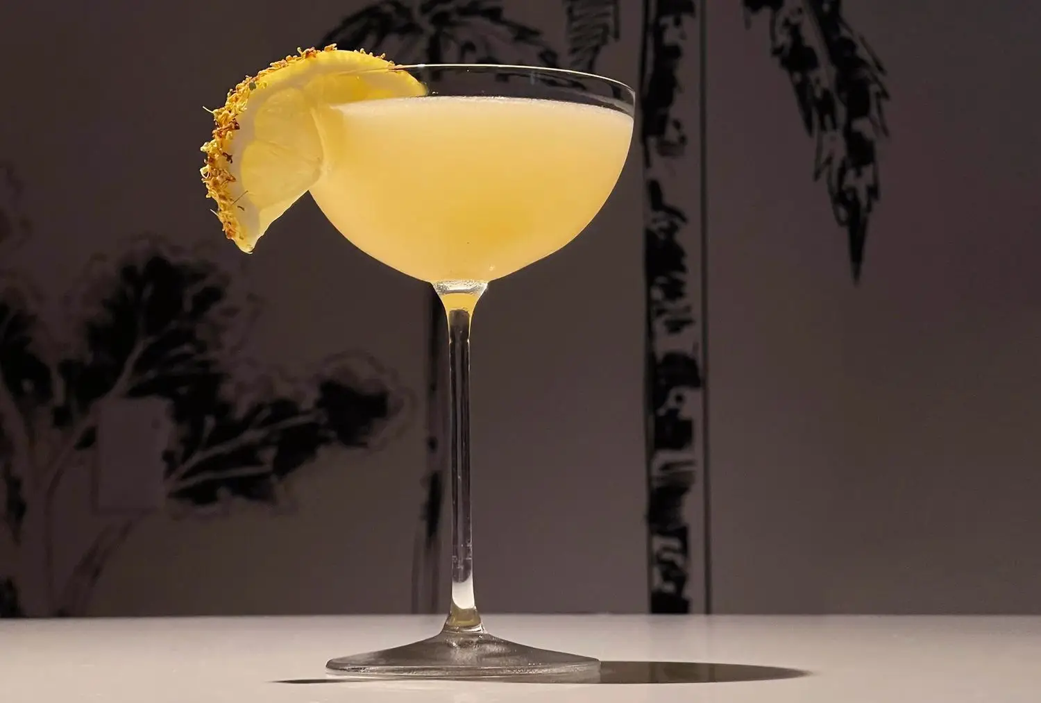 The Quynh Bee cocktail with spiced lemon garnish in coupe glass, on background showing printed tropical tree illustrations