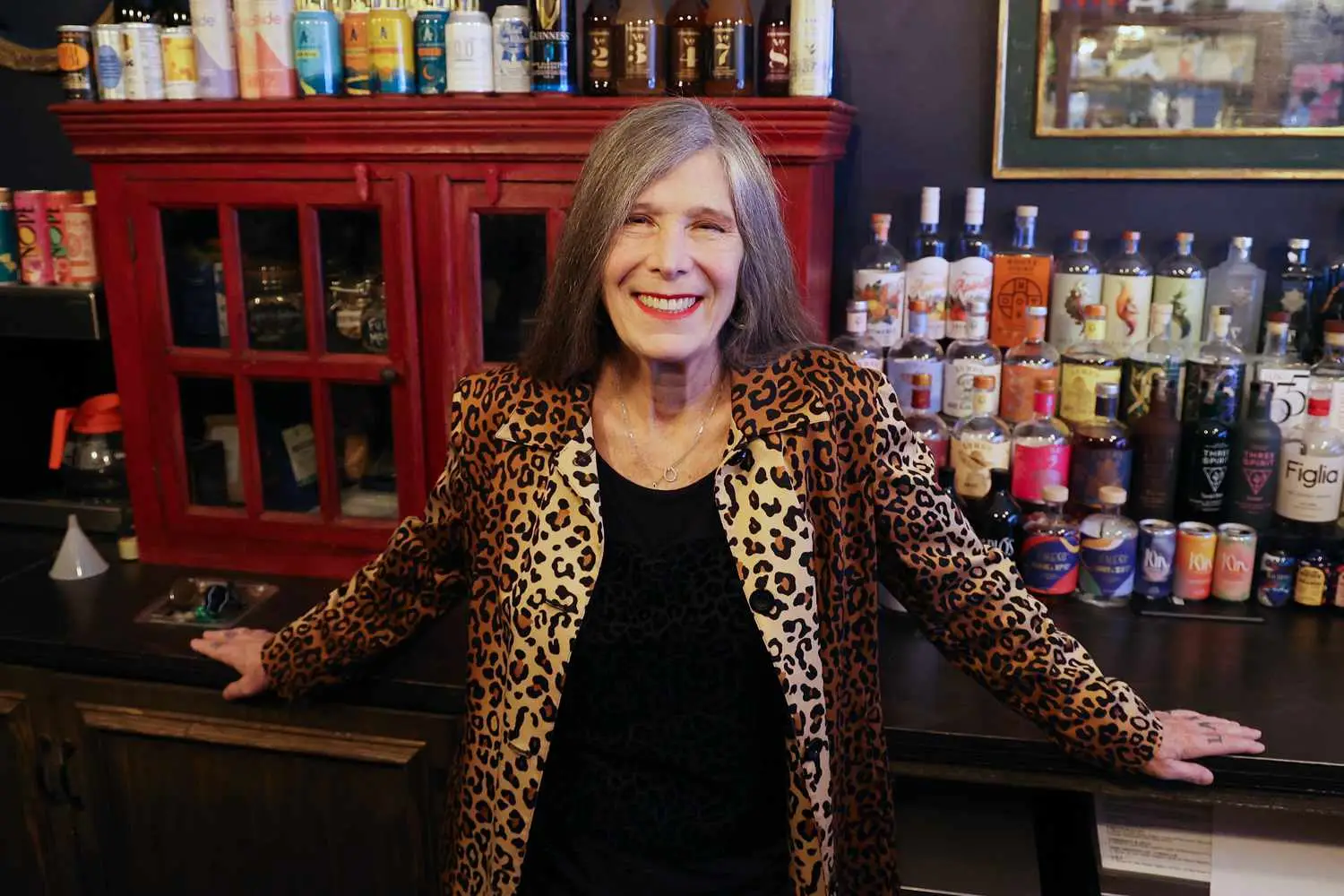 Abby Ehmann wearing leopard print jacket, standing behind the bar at Hekate in front of non-alcoholic bottles