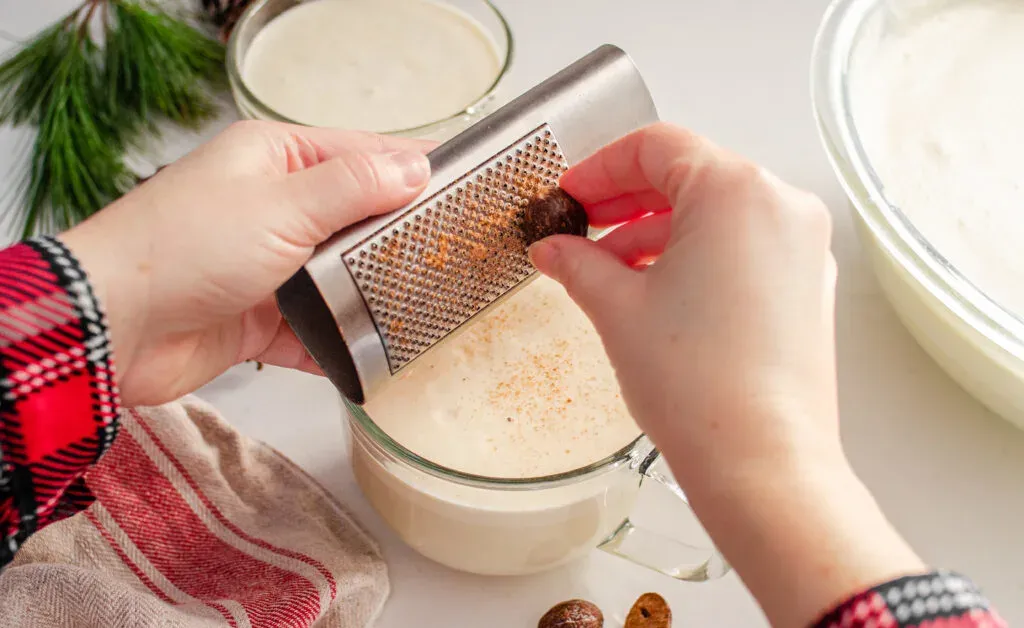 Grating fresh nutmeg over the mugs of my homemade cooked eggnog recipe without alcohol.' title='Homemade Eggnog