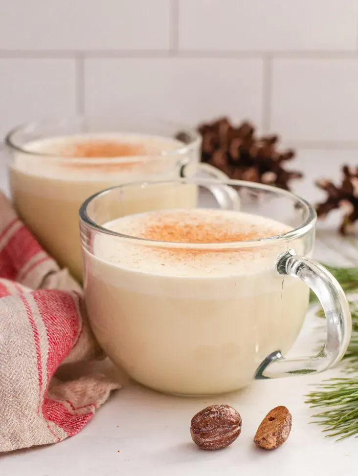 Two mugs of my homemade cooked eggnog recipe without alcohol with a tea towel greenery and fresh nutmeg around them.' title='Homemade Eggnog