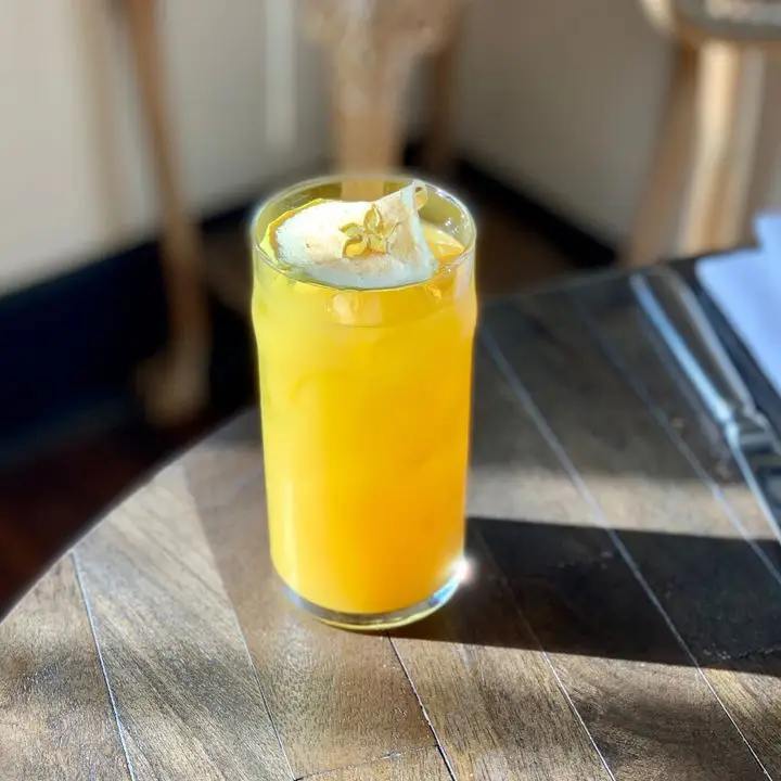 A golden yellow drink in a tall glass sits on a wooden table at Lou in Nashville. Topping the drink is a dried apple round, its center like a cut-out star