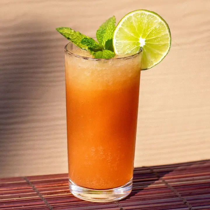 A collins glass rests on a bamboo mat. The drink within is bright, carrot-orange and blended with ice. It’s garnished with a lime wheel and mint sprig.