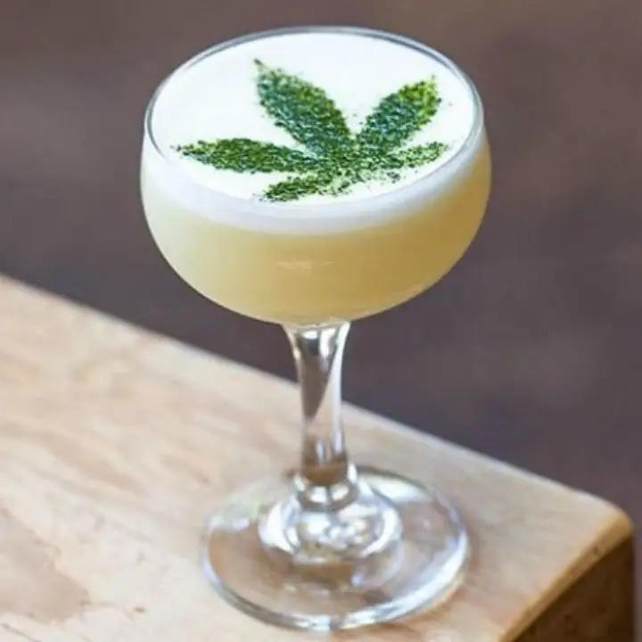 A rounded coupe rests on a pale wooden cutting board. The glass is filled with a hazy, pale gold drink topped with white foam; a cannabis leaf in matcha powder is stenciled onto the foam. 