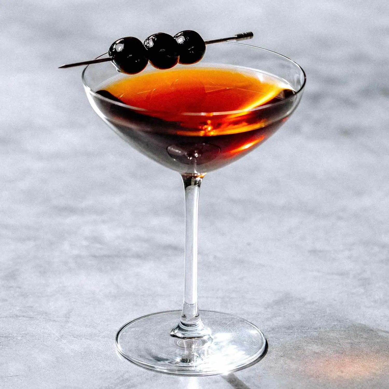Rob Roy cocktail in coupe glass with three brandied cherries, shot on gray marble surface