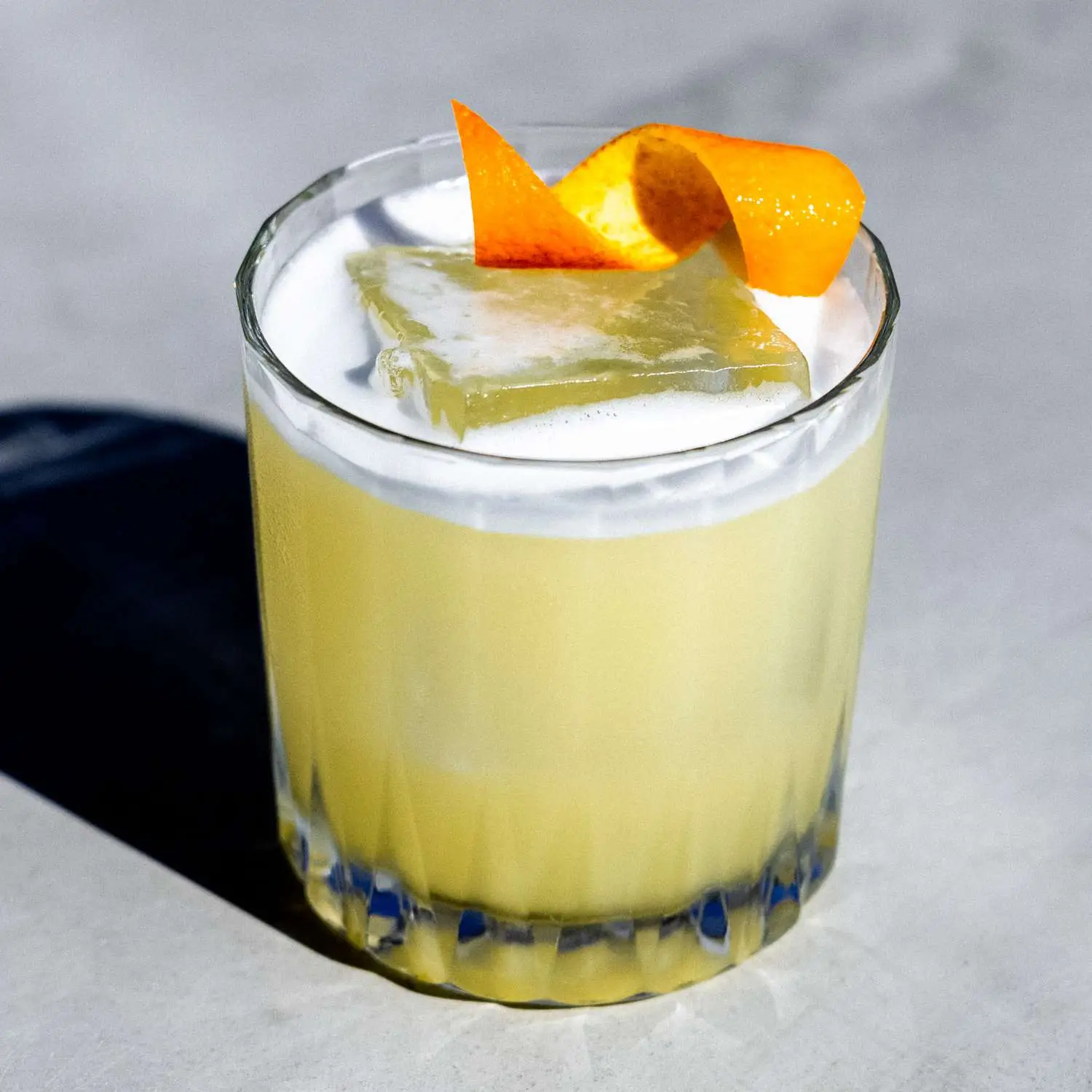 Scotch Sour with egg white foam on top, in a rocks glass with single large ice cube, and orange twist garnish
