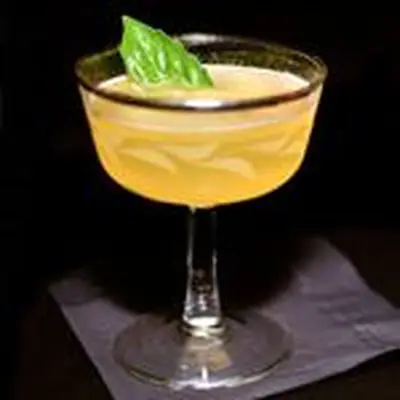 yellow-hued monastery cocktail in a gold-rimmed coupe, garnished with a basil leaf and served on a black napkin