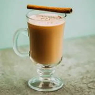 A glass mug holds a lush, golden-brown serving of Hot Buttered Rum. Its pale surface is flecked with nutmeg, and a cinnamon stick rests across the mouth of the mug. The background is pale blue, and the surface a light gray.