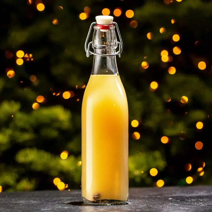 A golden, opaque beer fills a 250-milliliter stoppered bottle resting on a black marbled surface. The background is hazy and out of focus, with light coming through spruce trees.
