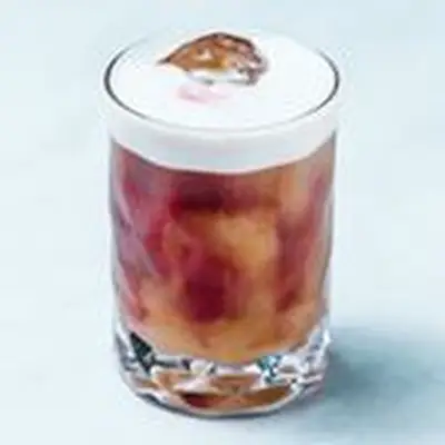 A tall, narrow rocks glass holds a vibrant drink, with red and gold colors swirling together around ice, topped with a thick white foam head. The background is a pale, off-white blue. 