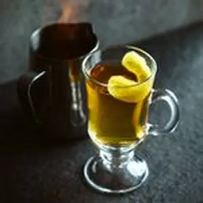 A clear glass cocktail mug filled with light brown liquid and garnished with a lemon twist; a metal pitcher with flames coming from within it sits to the left of the mug.