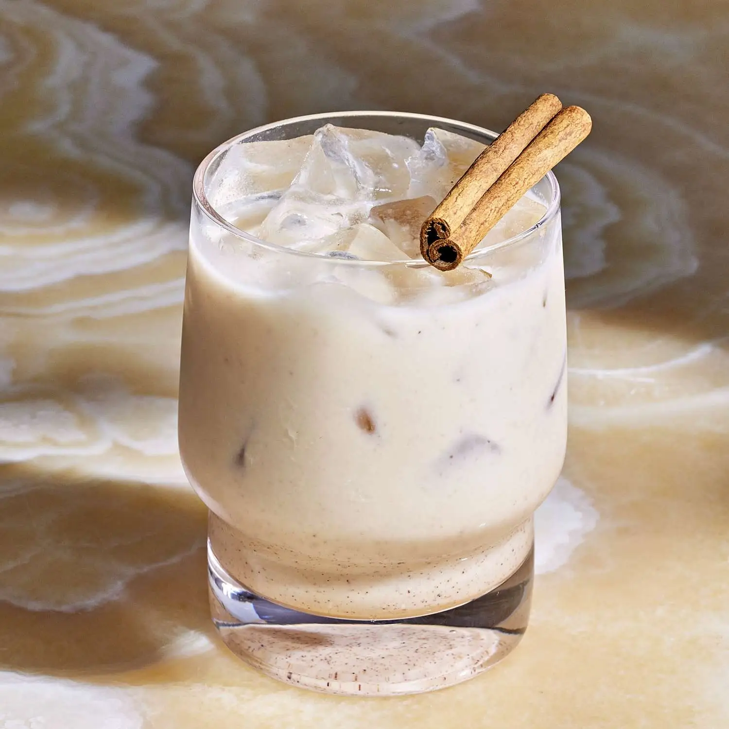Coquito in rocks glass on tan marble countertop, with cinnamon stick garnish