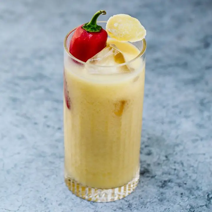 A Collins glass rests on a marbled stone surface. The glass is filled with ice cubes, a frothy yellow drink and a large red pepper. A candied piece of ginger rests atop.