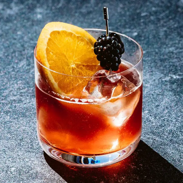 A thin-walled rocks glass rests on a black marble surface. It’s filled with a few ice cubes, a red-orange drink, an orange wheel and a blackberry on a silver pick.