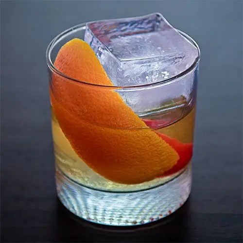 A light amber cocktail on ice in a rocks glass garnished with orange zest and a Fresno chile