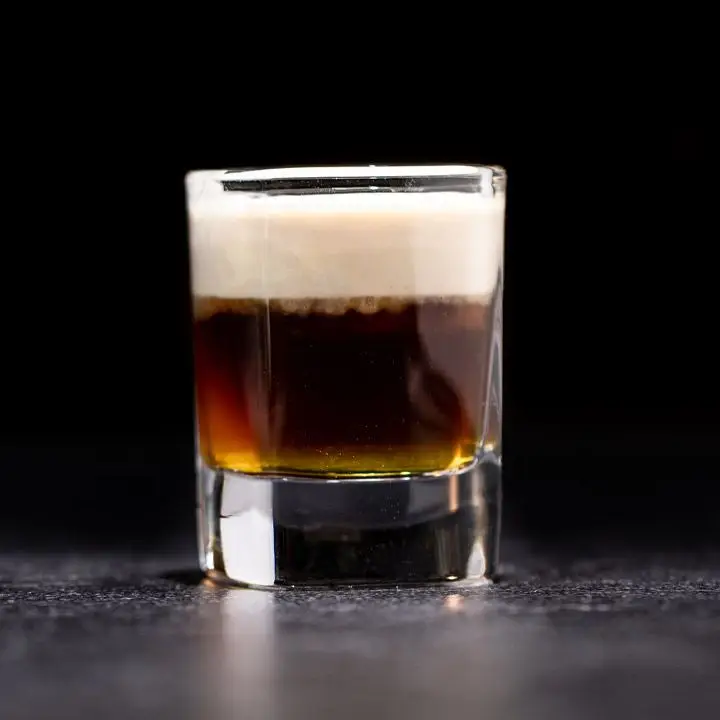 A squat shot glass sits on a black and gray surface, the background is dramatically black. The shot glass holds a layered drink of yellow Galliano, dark brown espresso and a float of white cream.