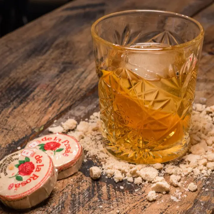 A rocks glass with a star insignia etched in the glass sits on a wooden table. It’s filled with a large ice cube, a pale gold drink, and an orange peel. Around it is crumbled marzipan and two De La Rosa candies.