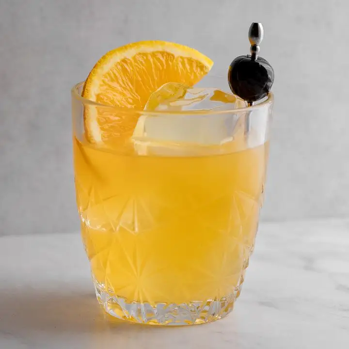 A rocks glass with star shapes holds a bright orange cocktail with a few large ice cubes. It’s garnished with an orange slice and a cherry on a silver pick.