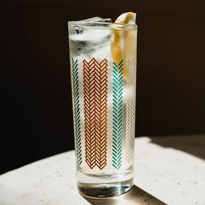 A highball glass sits on a dramatically lit stone surface. It is marked with red and blue chevrons, and is filled with a clear sparkling drink, ice and a lemon wedge.