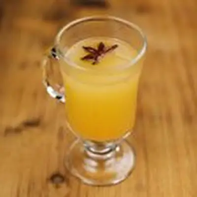 A glass mug with a handle and stem sits on a wooden surface. It’s filled with an opaque golden drink and garnished with a star anise.