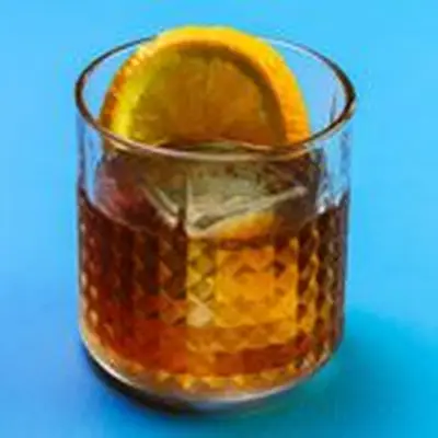 On a sold blue backdrop, a multi-faceted rocks glass holds a single large ice cube, a full orange wheel, and an amber-colored bourbon drink. 