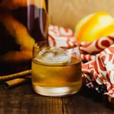 A short, wide rocks glass with thin walls rests on a dark wood surface. It’s filled with dark whiskey and a large ice cube, and surrounded by a red and white cloth, cinnamon sticks, a jar and a partially peeled lemon.