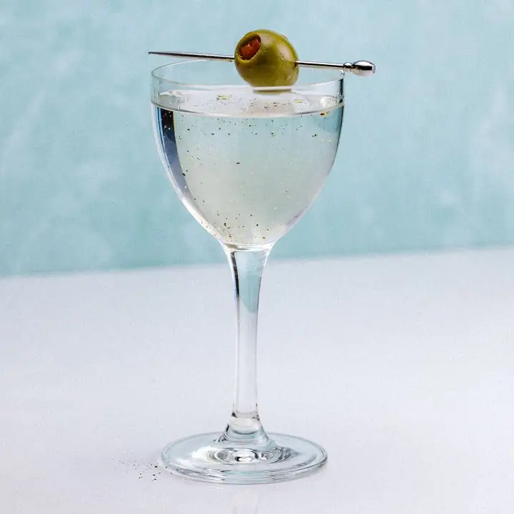 On a white counter with a sky-blue background, a beautiful curved Nick & Nora glass holds a Martini. The drink is slightly cloudy and decked with spots of pepper. A single pepper-stuffed olive is pierced on a silver skewer resting across the mouth of the glass.