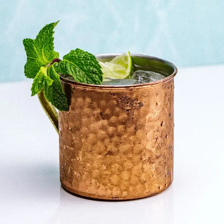 A hammered-copper Moscow Mule mug sits on a white surface. A large sprig of mint drapes over the mouth of the glass, with a lime wedge behind it. The background is minimal, and pale blue.