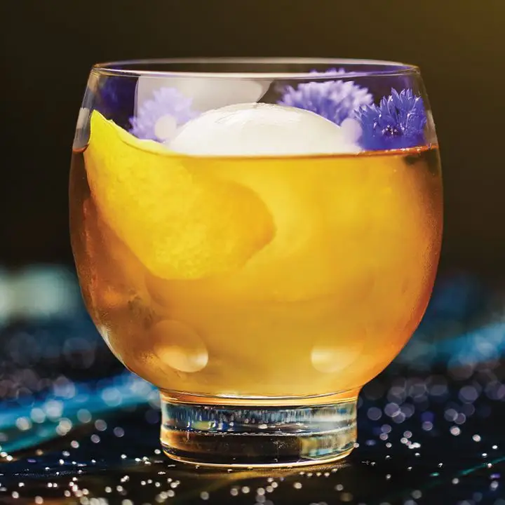 Guinan’s Negroni cocktail in a round-bowled double rocks glass, served over an ice sphere and garnished with a lemon peel and purple flowers
