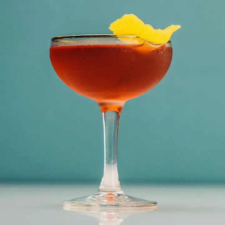 Old Pal cocktail in gold rimmed coupe glass with lemon peel garnish, shot against blue wall