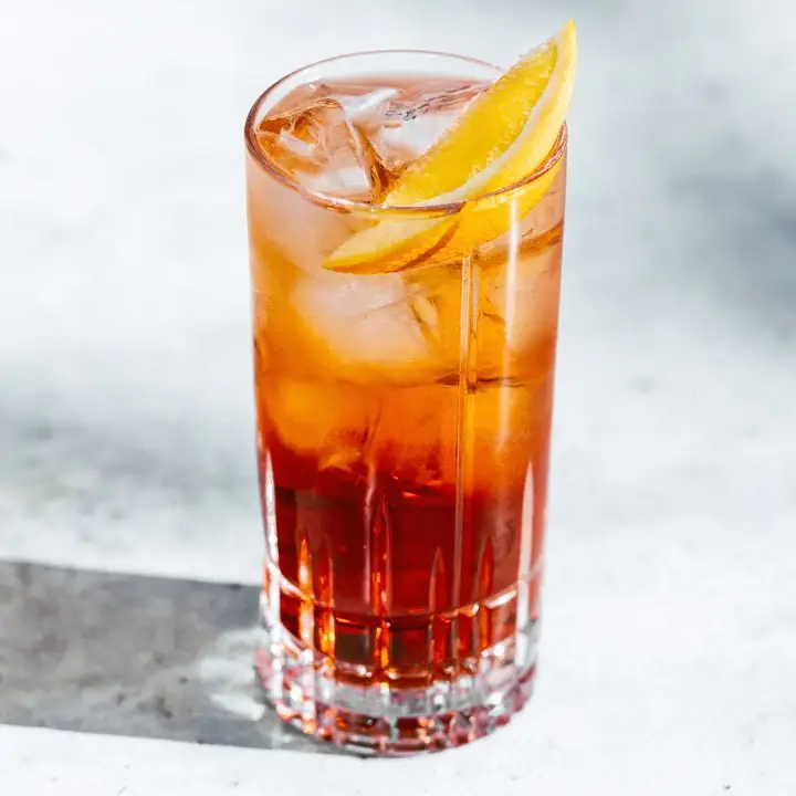 Americano cocktail in highball glass with ice and orange wedge, shot on white-gray marble