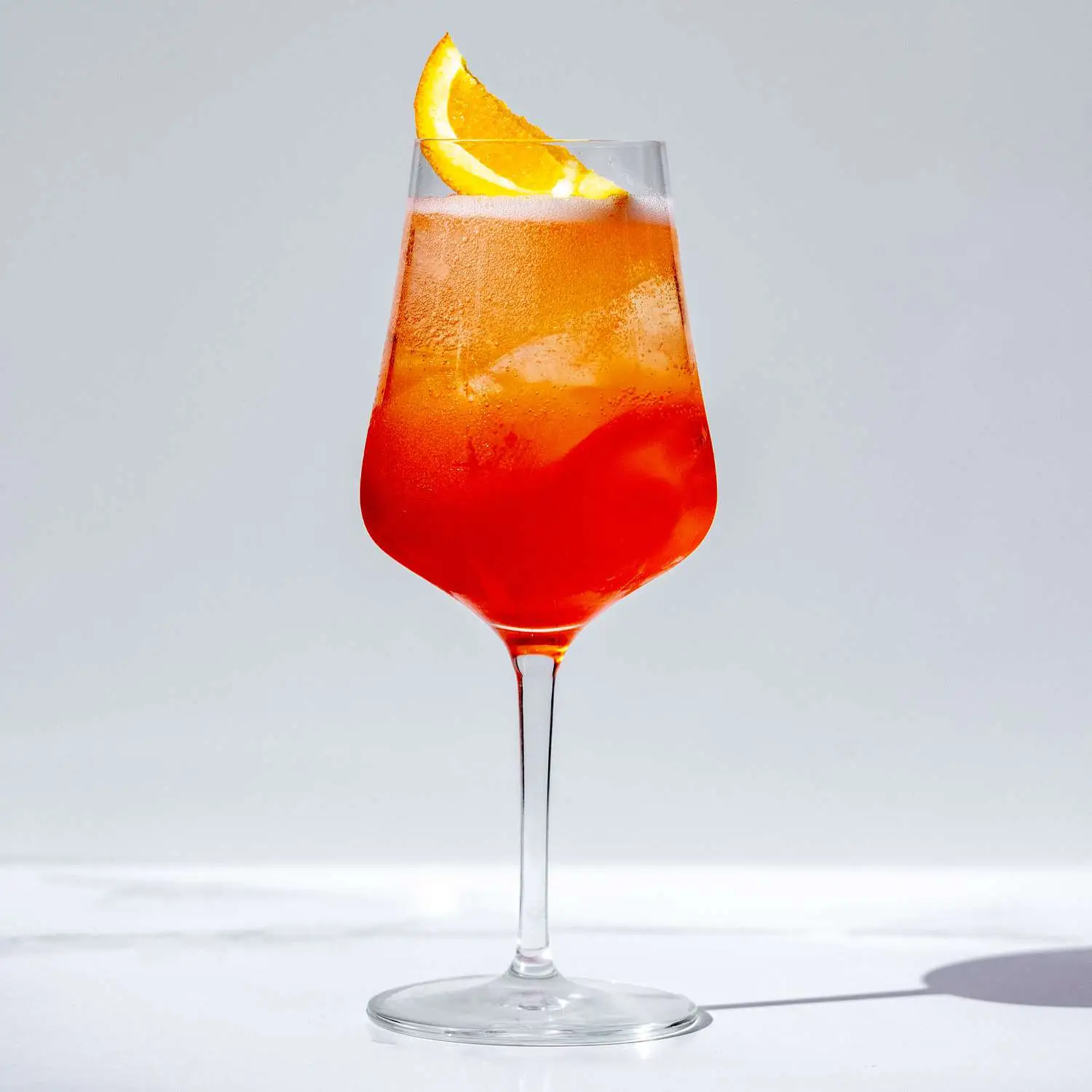 Vibrant Campari Spritz cocktail in wine goblet with ice and orange wedge, shot against white