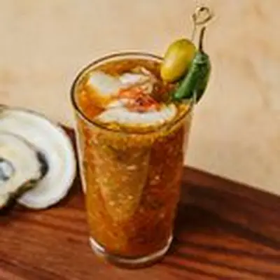 Bloody Roman cocktail in a pint glass, topped with two oysters, a green olive, serrano pepper and red chile threads