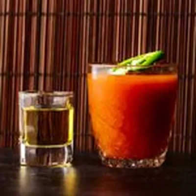 Two glasses rest on a black surface in front of a bamboo mat. On the left is a squat shot glass of tequila. On the right is a larger glass of vivid orange Sangrita, garnished with a jalapeño.