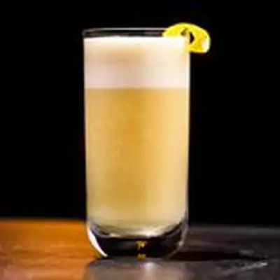 A clear, straight walled highball glass is filled with a foamy, bubbly copper-colored drink topped with a thick layer of foam. It’s garnished with a small lemon peel. The background of the photo is black, and the drink is dramatically lit, resting on a bar top. 