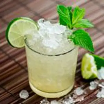 Crushed ice and pale spirits fill a short rocks glass, which rests on a wooden curtain. Ice and lime spill out of the cup, and the drink is garnished with mint and a lime wheel.