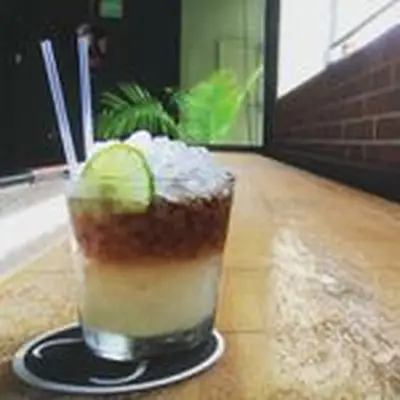 On a long bar, a short rocks glass is filled with crushed ice. The drink includes two layers: a top dark brown layer, and a bottom light yellow. Built over crushed ice, it’s garnished with two straws and a lime wheel.