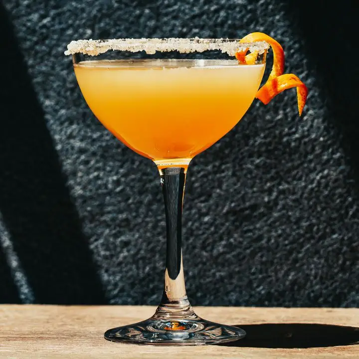 A curved martini glass rests on a wooden surface in front of a black stone wall. The glass is filled with an orange-colored cocktail and garnished with a spiral twist of orange. The rim is covered in sugar.