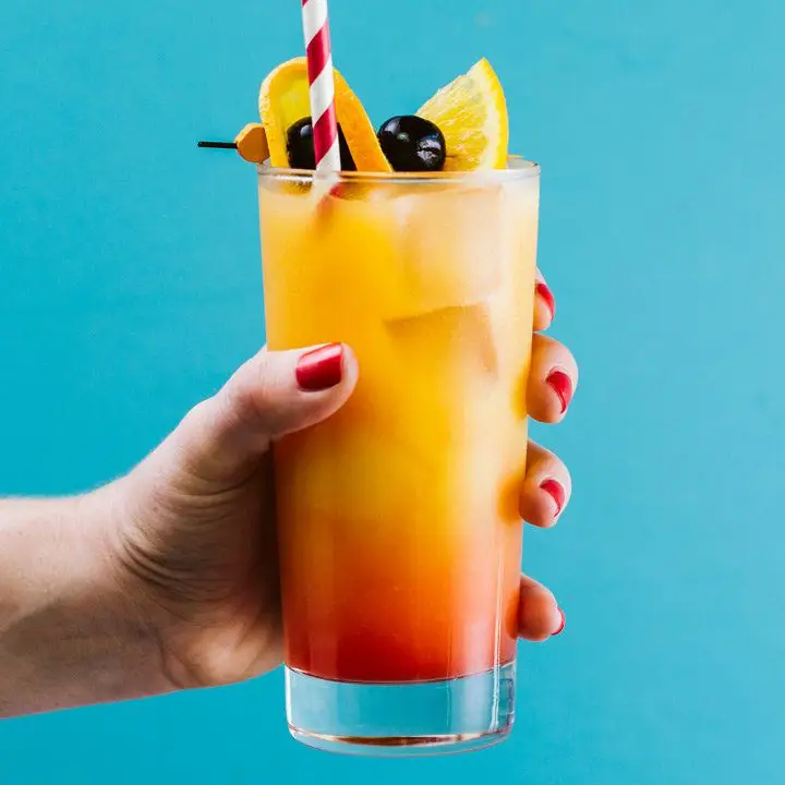Hand holding Tequila Sunrise in highball glass with straw, garnished with two cherries and an orange, shot against blue background