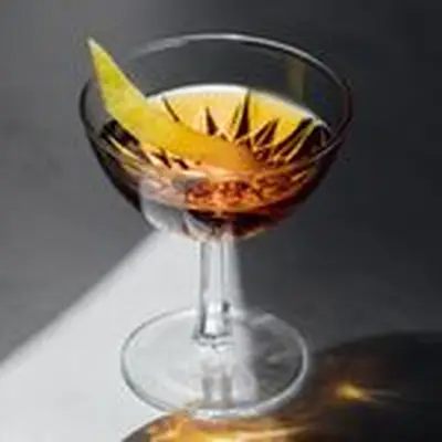 A faceted cocktail coupe sits on a marble surface. A beam of light illuminates a golden beverage within, and a thin slice of lemon peel. The rest of the photo is in shadow.