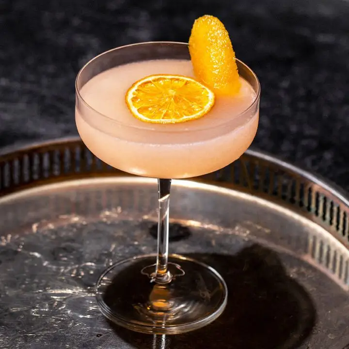 A flat coupe glass is filled with a pink-orange Cosmopolitan variation. A dehydrated orange wheel floats in the center of the drink, while an orange peel pokes up as a garnish. The drink is presented on a silver platter with a tall rim. 