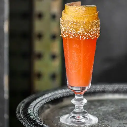 A tall, narrow crusta glass holds a red orange drink. A curled grapefruit peel rests in the mouth of the glass, which is dusted with sesame seeds.