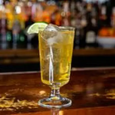 A stemmed Collins glass holds a golden yellow sparkling drink over ice cubes. It sits on a wooden bar, and is garnished with a lime wedge.