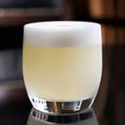 xA light yellow, frothy Pisco Sour in a rocks glass on a black reflective surface