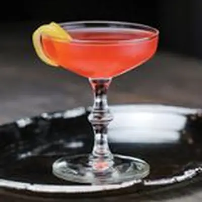 An elegant coupe glass holds a bright red drink with a thin slice of lemon peel. The glass sits on a silver tray 